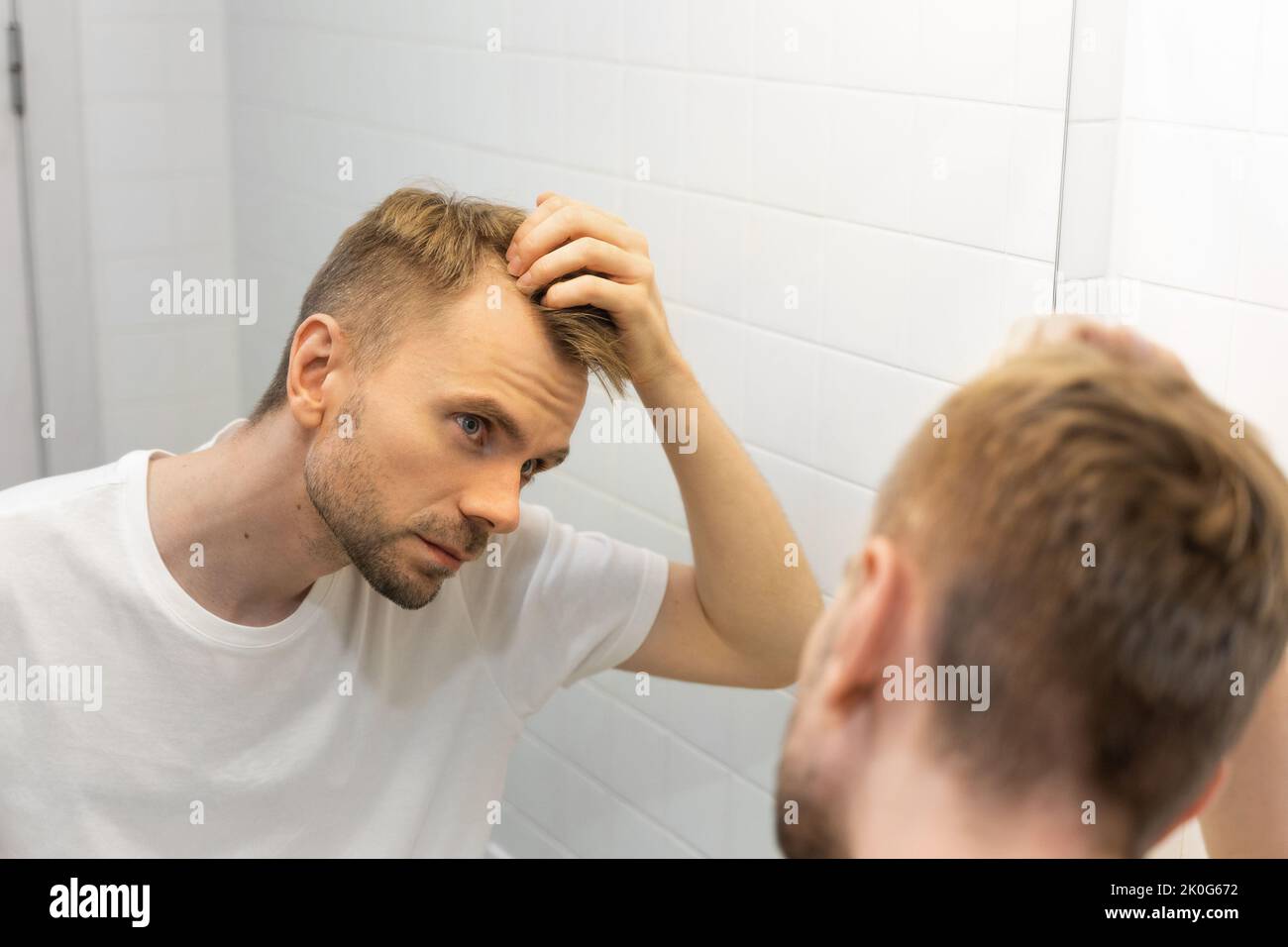 Middle aged caucasian white man with a short beard looks at his hair in the mirror in the bathroom and worried about balding. Concept of hair problem Stock Photo