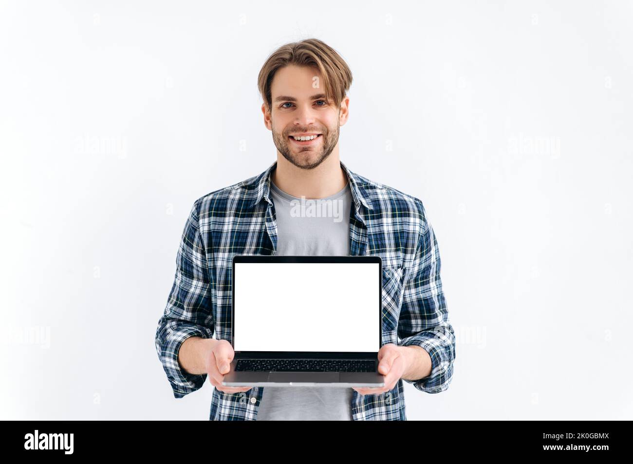 Mock-up concept. Handsome modern caucasian guy in casual wear, freelancer, student, holding an open laptop with empty mock-up space, stand on white isolated background, looks at camera, smiles Stock Photo