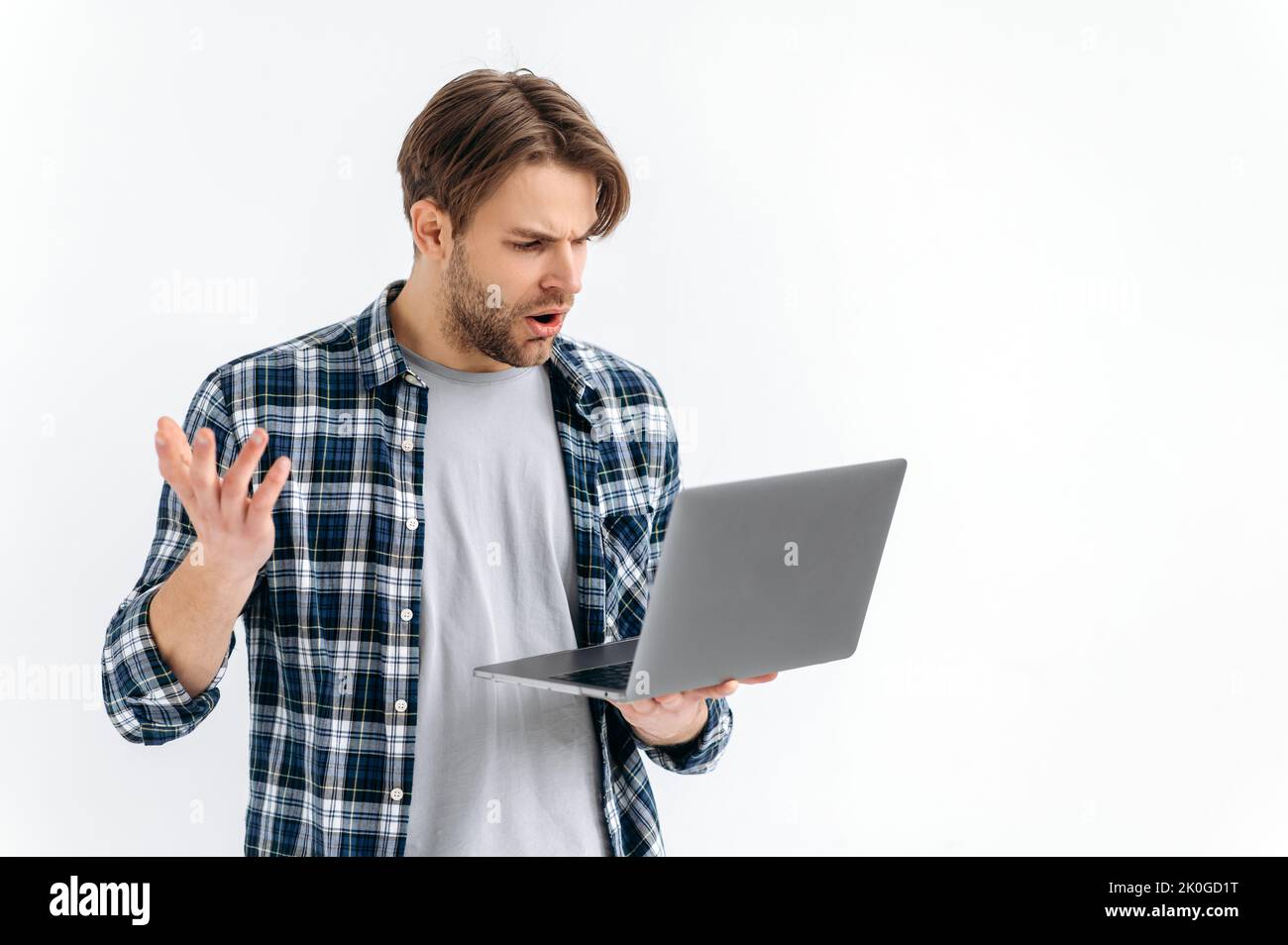 Frustrated confused puzzled caucasian stylish young man, standing on a white isolated background, holds an open laptop, surprised looks at the screen, see unexpected news, message, gesturing with hand Stock Photo