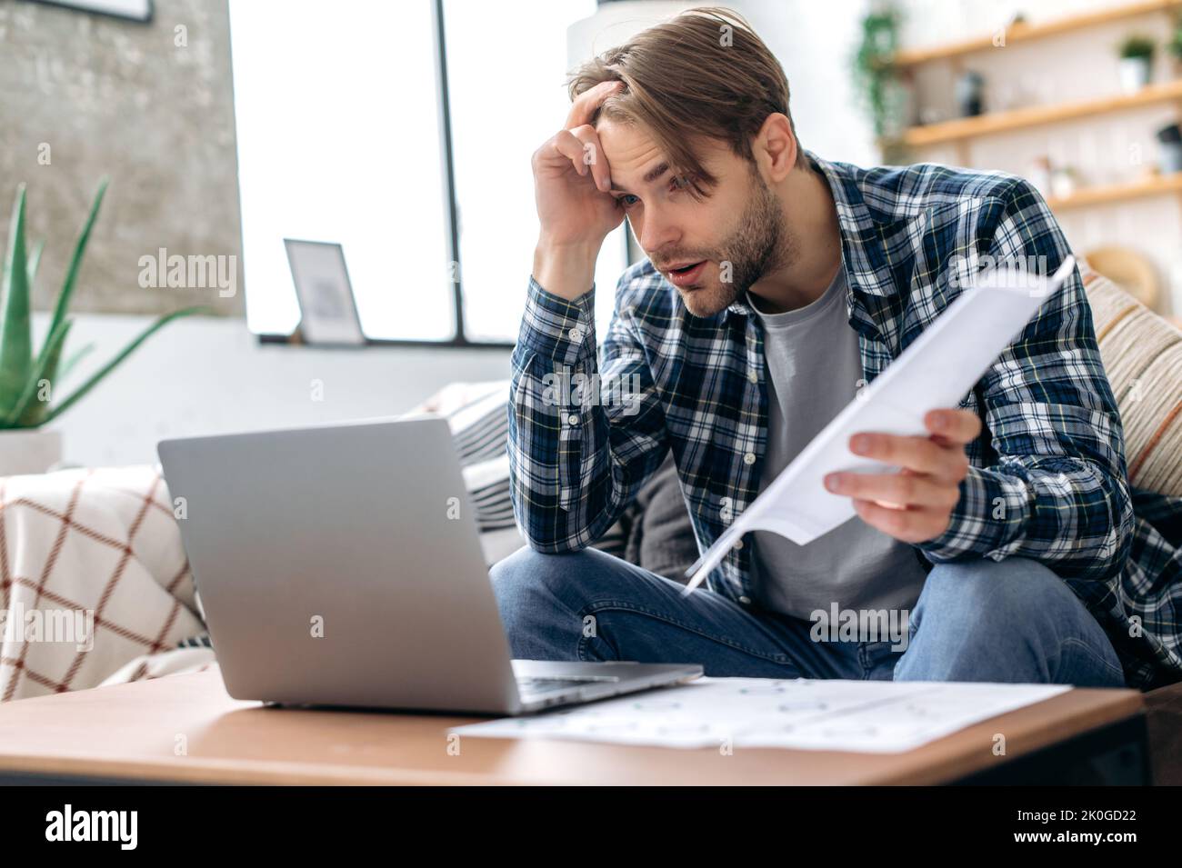 Frustrated sad caucasian young man in stylish casual clothes, freelancer, web designer, ceo, sitting in living room on the sofa, working on a laptop on a project, analyze documents, upset with result Stock Photo