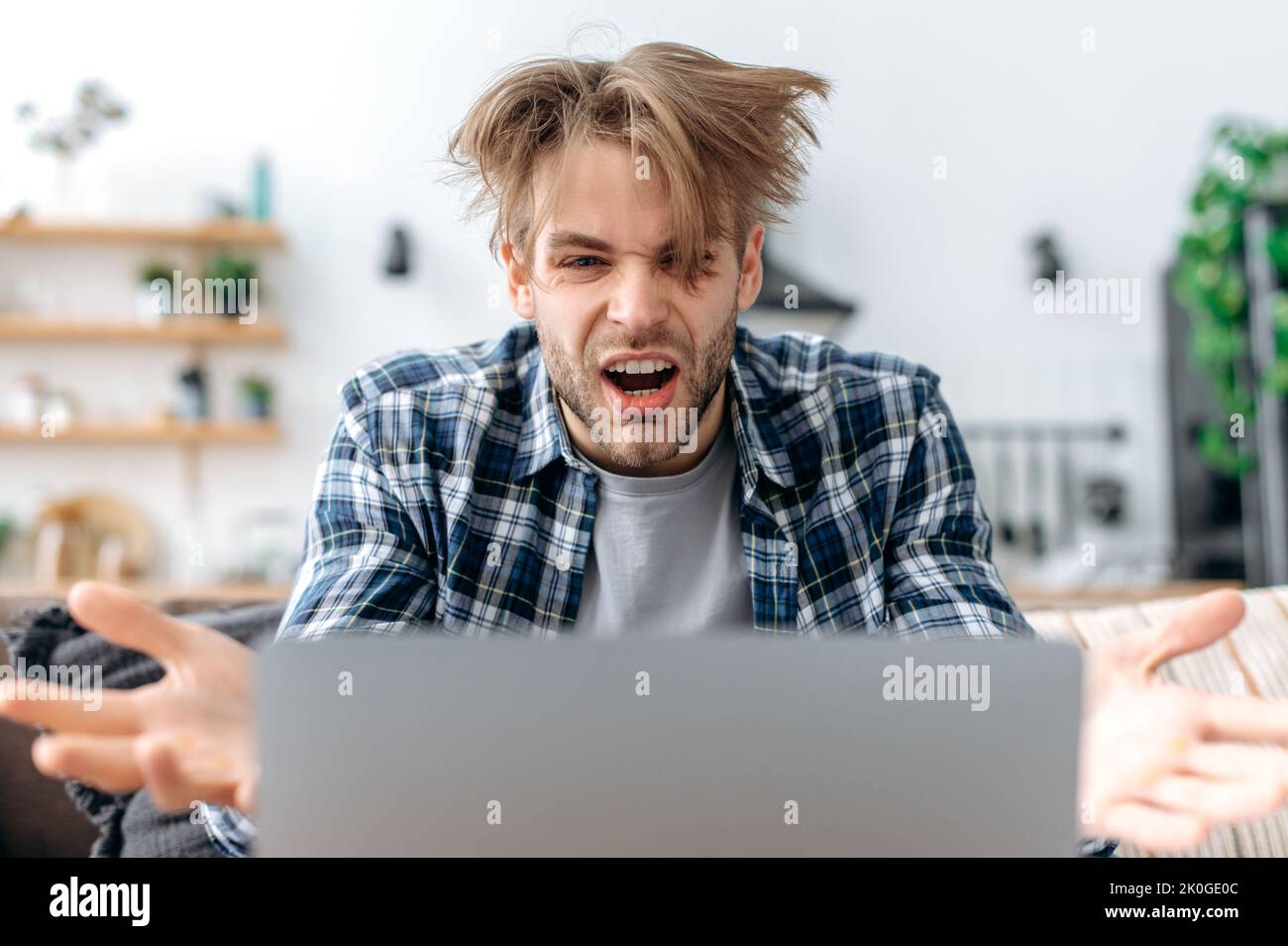 Angry annoyed caucasian young man in stylish casual clothes, freelancer, web designer, ceo, sitting in living room, working on a laptop on a project, yelling at a screen, irritated with result Stock Photo