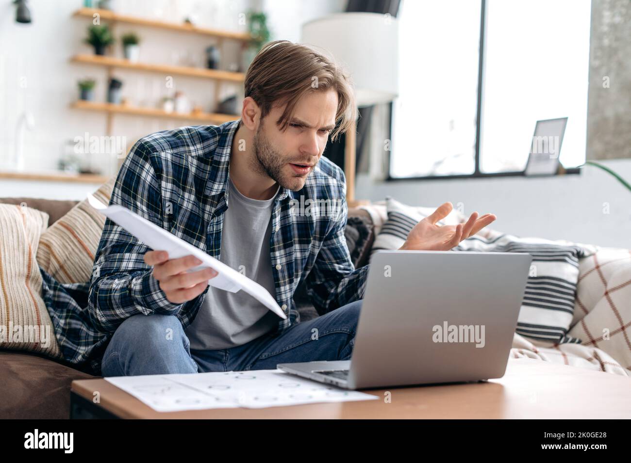 Puzzled confused caucasian young man in stylish casual clothes, freelancer, web designer, sitting in living room on the sofa, working on a laptop on a project, analyze documents, misunderstanding Stock Photo