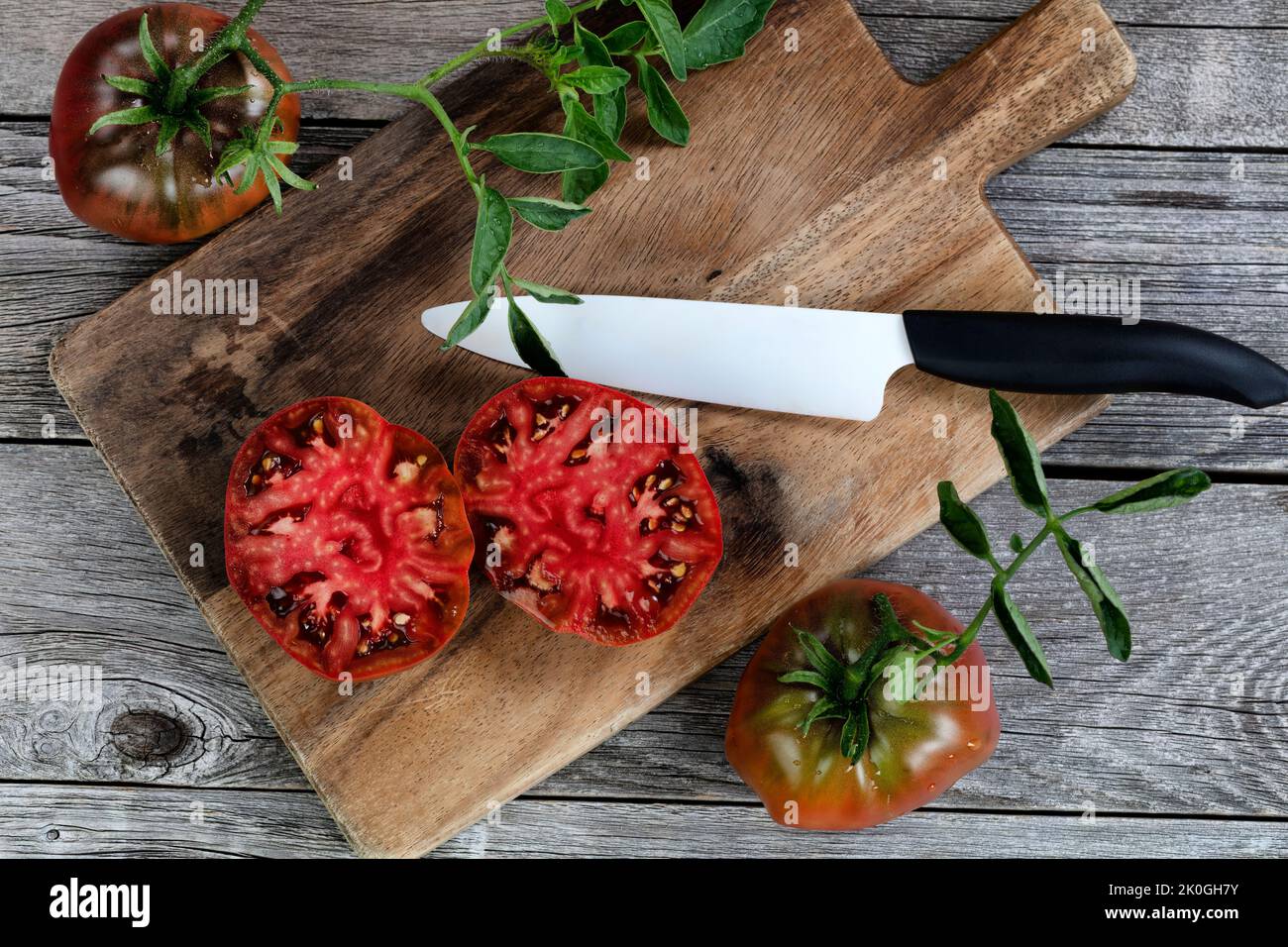 Ripe organic tomato sliced in half with cutting ceramic knife plus server on rustic wood table in flat lay format Stock Photo