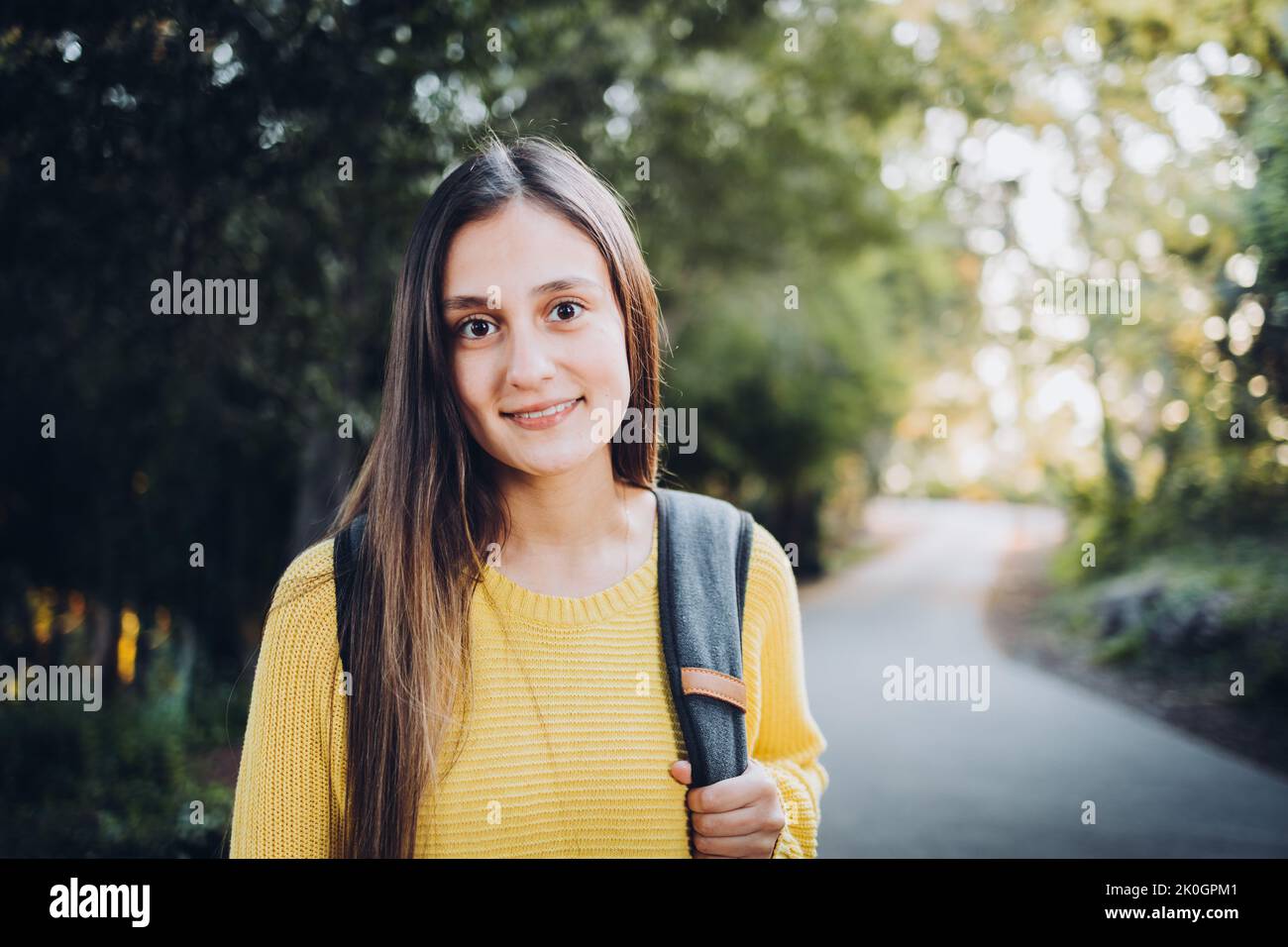 Smiling university student woman wearing a yellow sweater and carrying a backpack in the campus park Stock Photo