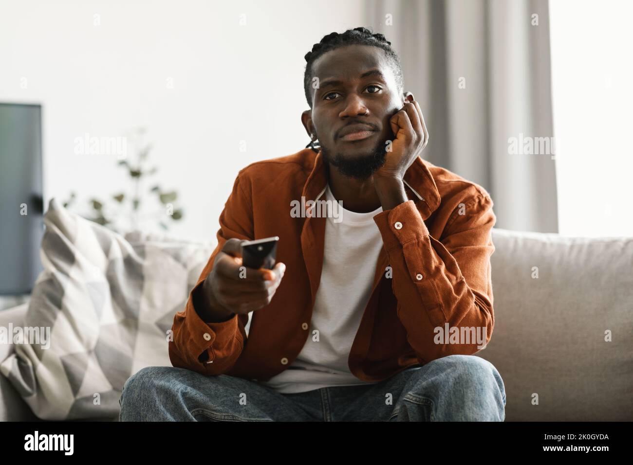 Boring television program. Bored upset black man watching TV and switching channels with remote control, sitting on sofa Stock Photo