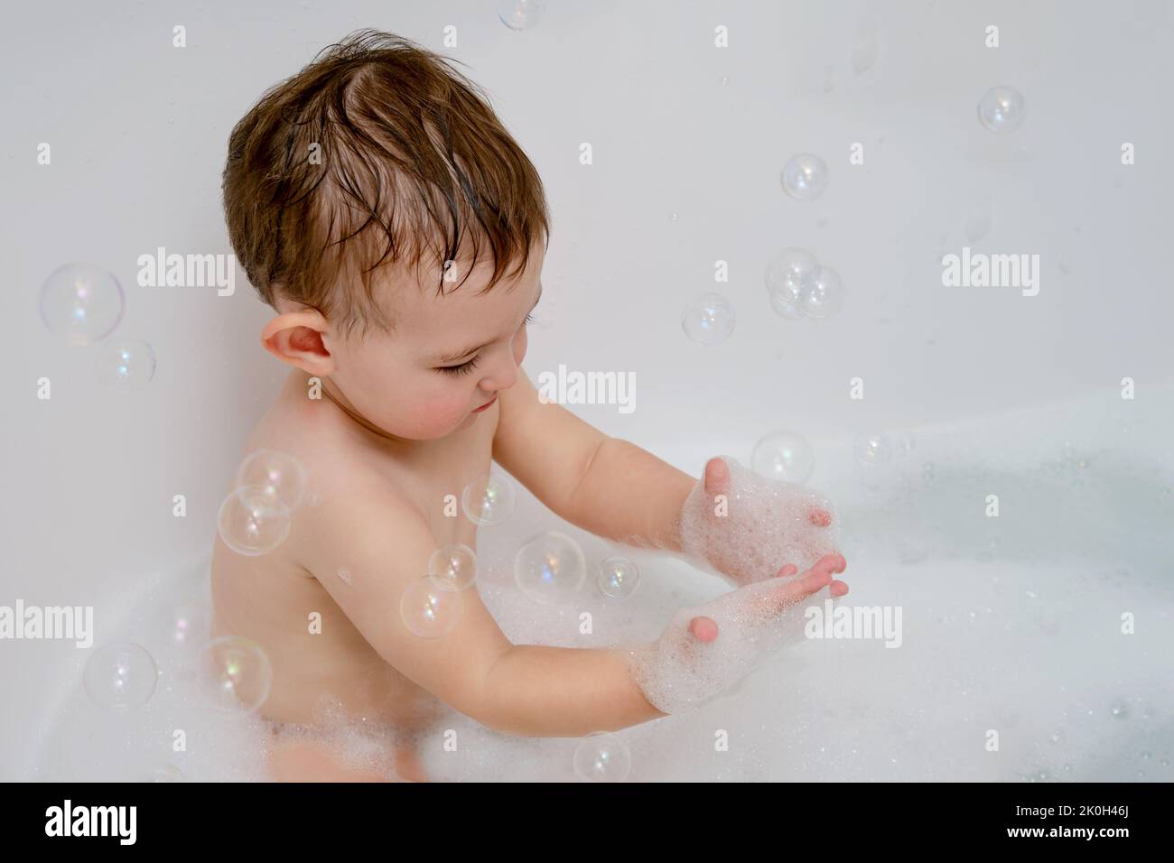 Happy toddler baby is playing in a white bathtub with soap bubbles
