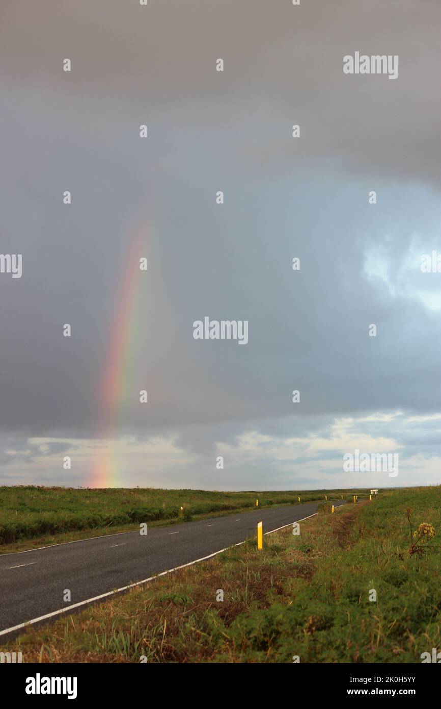 A vertical shot of an asphalt road with a rainbow above it during daytime in Iceland Stock Photo