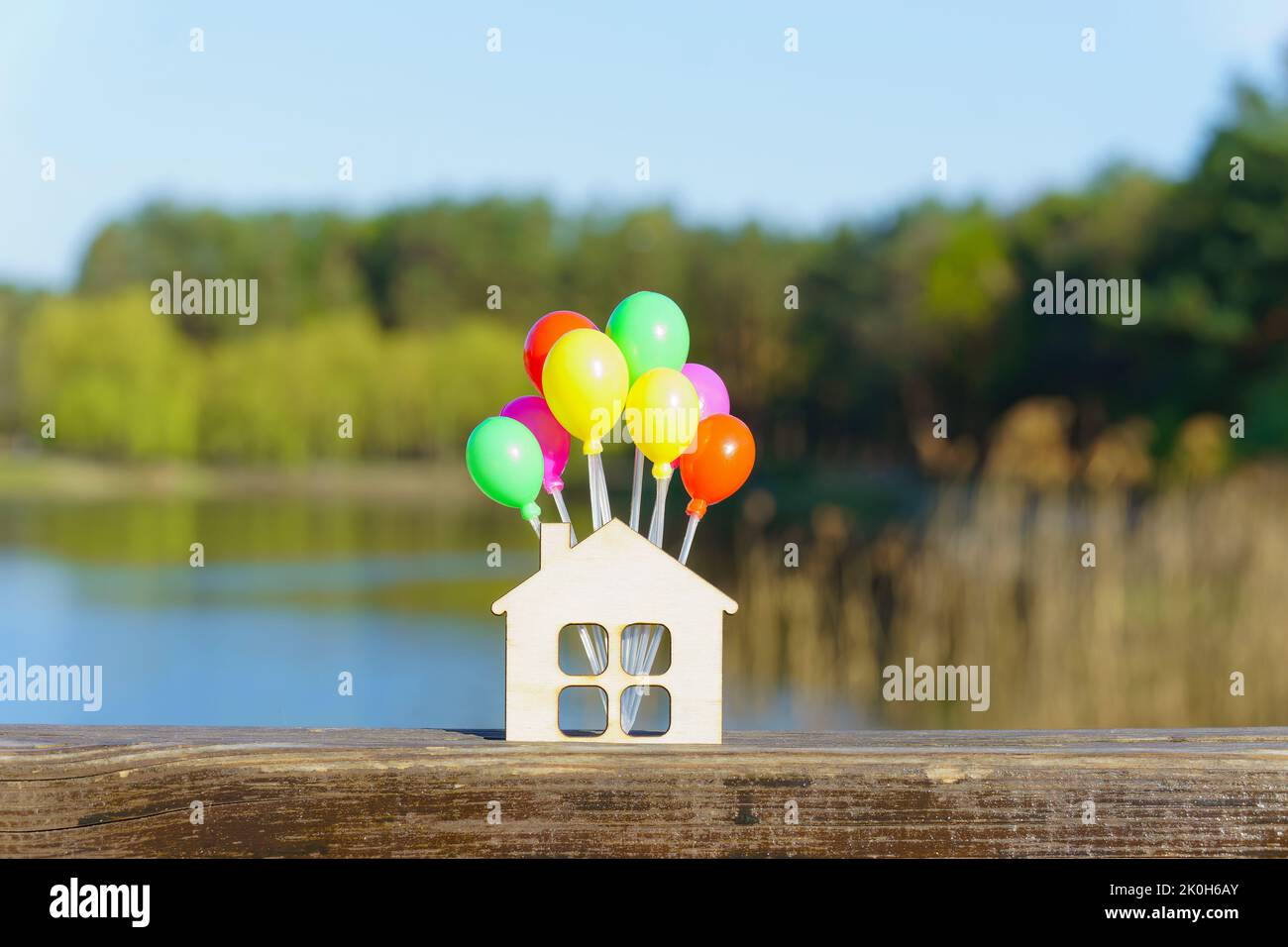 Flat wooden house shape with a bunch of colorful balloon props against a beautiful lake background. Buying a countryside house. Stock Photo