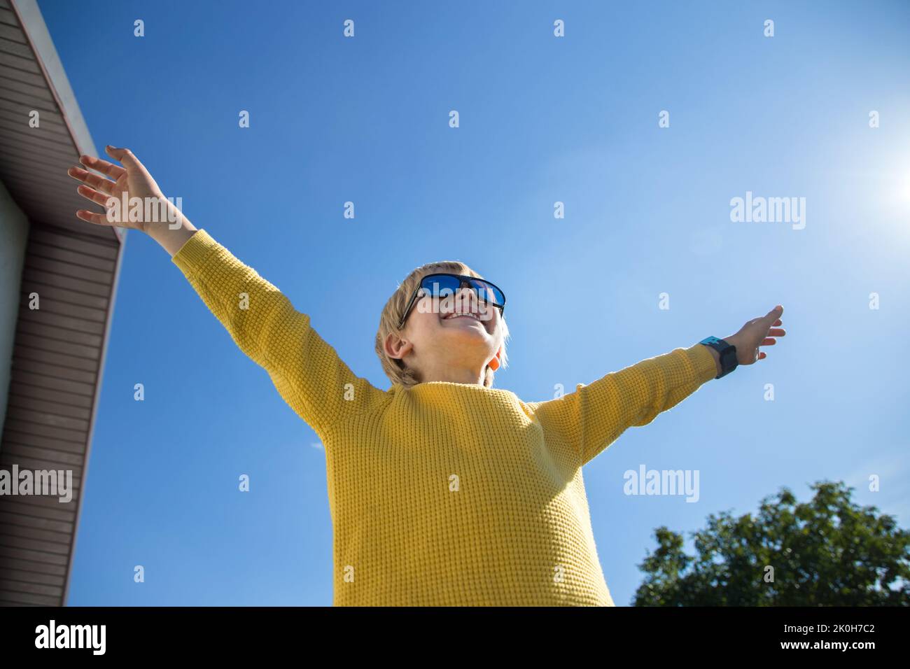 joyful boy in yellow sweater, sunglasses spread his arms against of blue sky and sun. victory, success, joyful emotions, happiness, freedom, positive Stock Photo