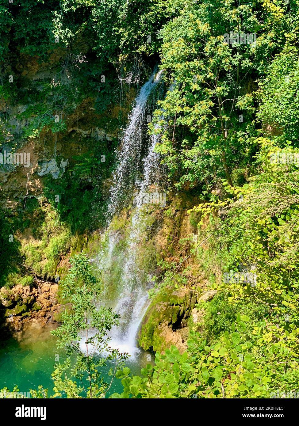 A vertical shot of a beautiful small waterfall in a forest in sunlight ...