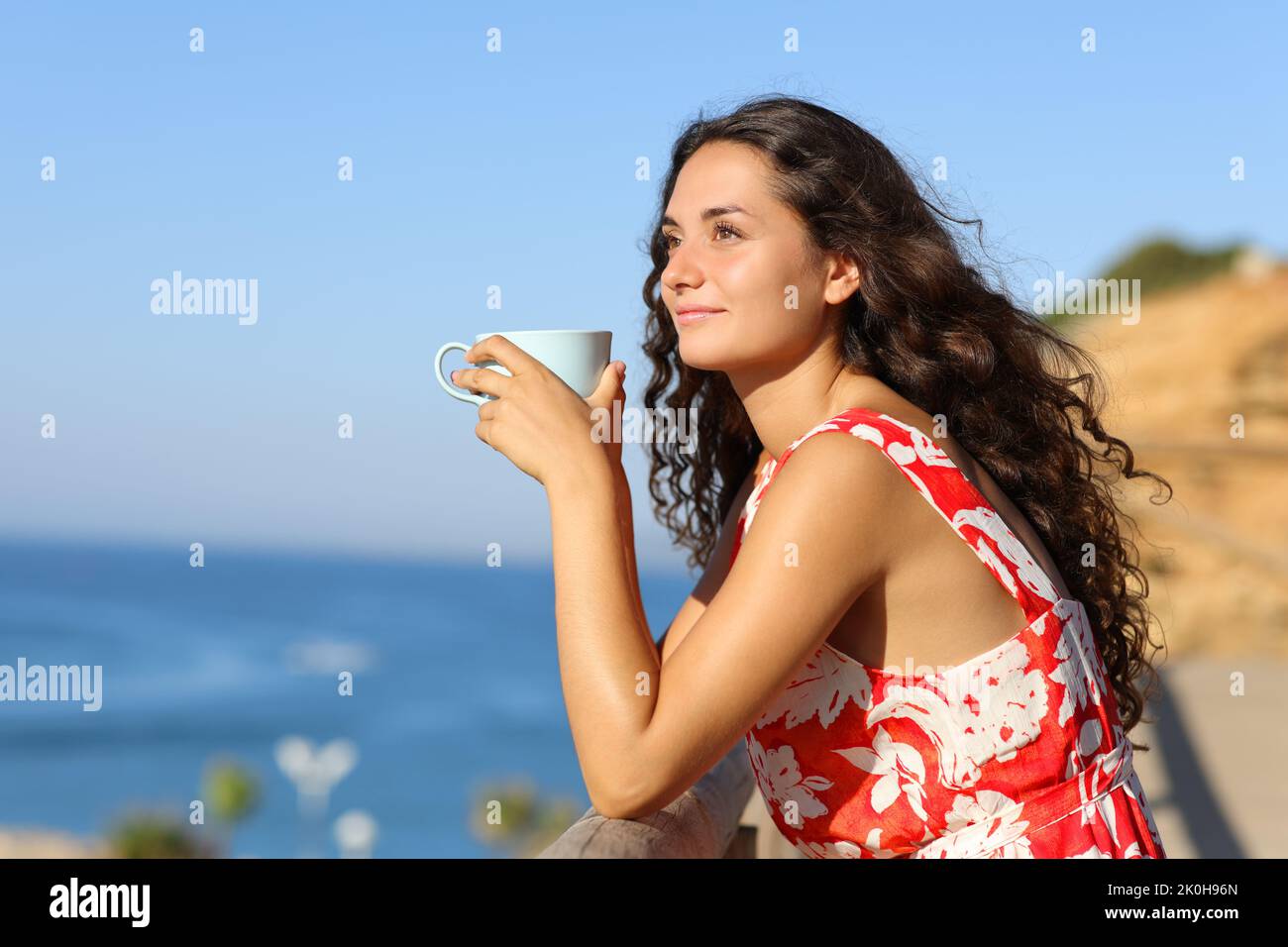 Woman in red drinking coffee on the beach looking away a sunny day Stock Photo