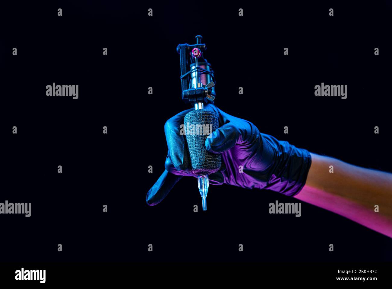 Closeup of tattooer master's hand in black glove holding machine for making tattoo art on body isolated on dark background in neon light. Stock Photo