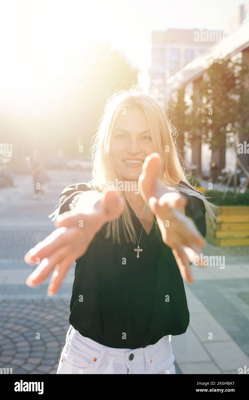 Sunlit portrait of an attractive young blond woman reaching with her hands, looking at the camera. She is standing on a pavement on a street. Stock Photo