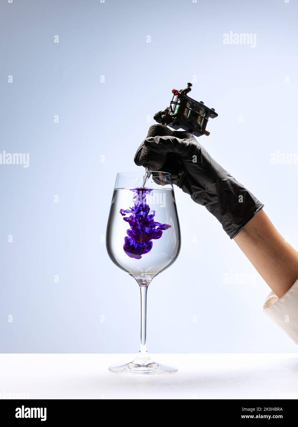 Wine glass and purple ink. Tattooer master's hand in black glove holding machine for making tattoo art on body isolated on white background Stock Photo