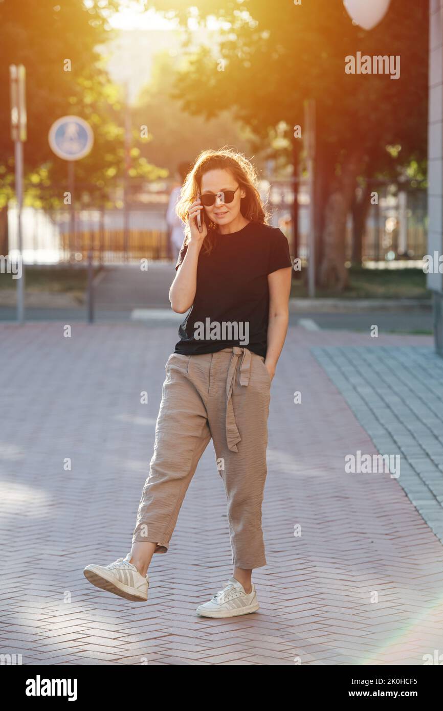 Strolling young woman in sunglasses listening to her phone while leisurely walking on a pavement on a sunlit street. Stock Photo