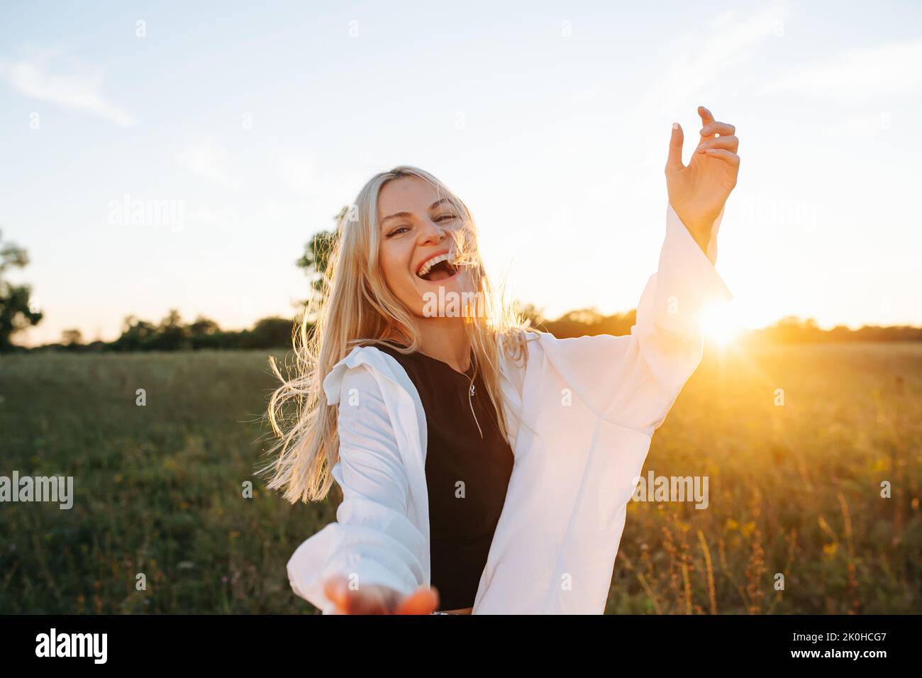 Cheerful young blond woman standing amidst wheat field. Reaching with her hand. Against setting sun, lit with soft bright orange light. Low angle. Stock Photo