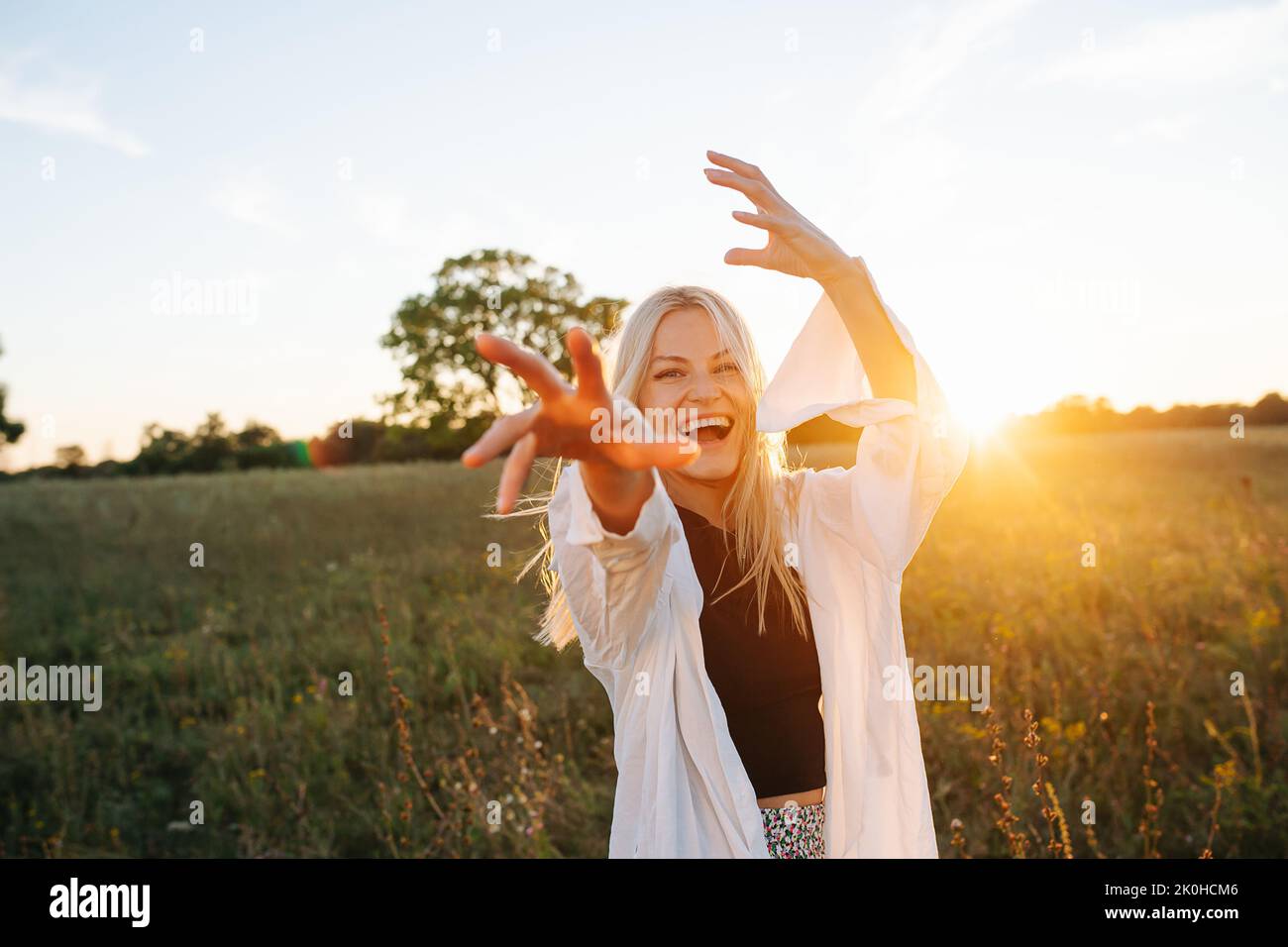Overjoyed young blond woman standing amidst wheat field. Reaching with her hand. Against setting sun, lit with soft bright orange light. Low angle. Stock Photo