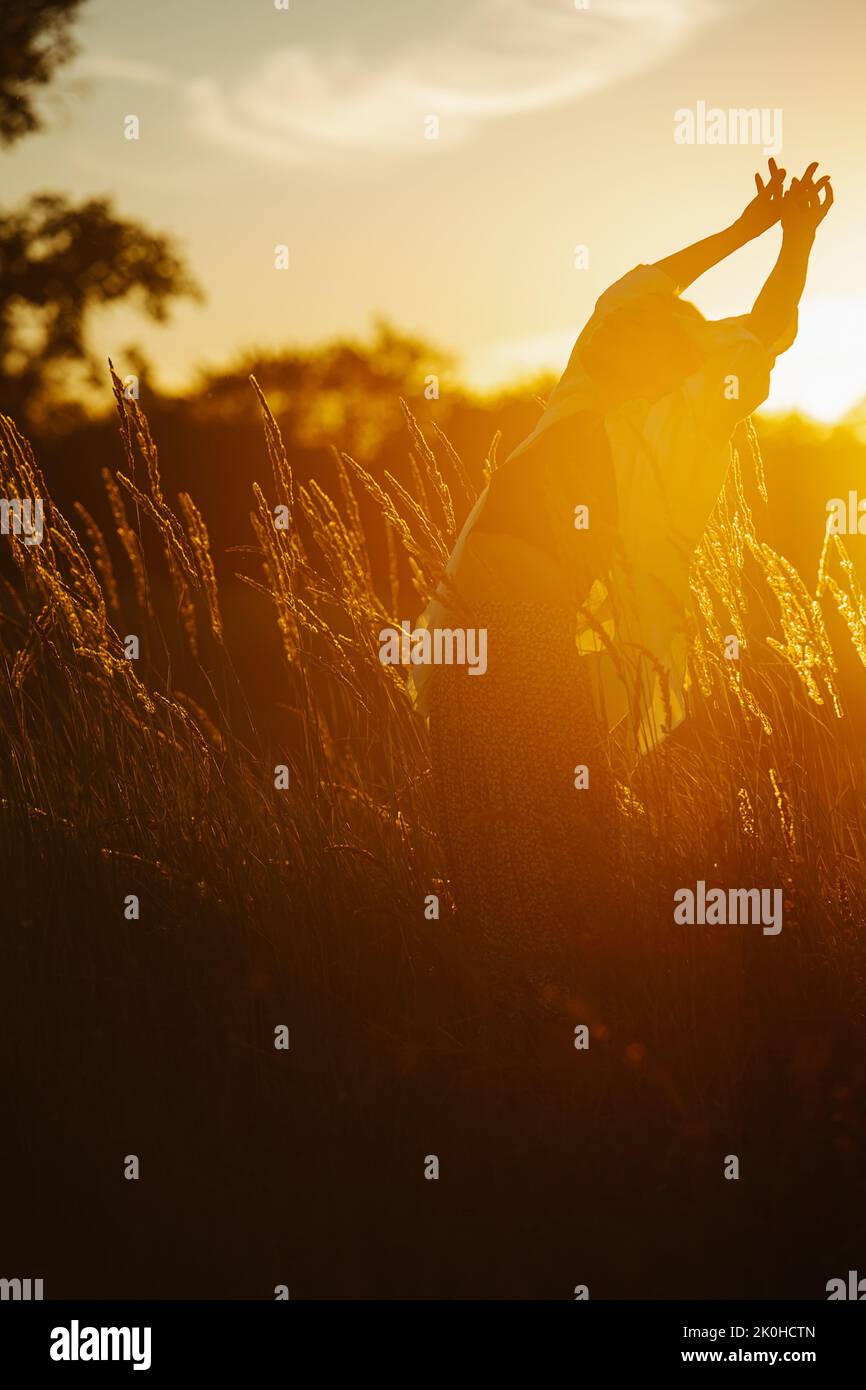 Stretching, bending back young blond woman standing amidst wheat field. Low angle. Dark shape against blinding light of a setting sun Stock Photo