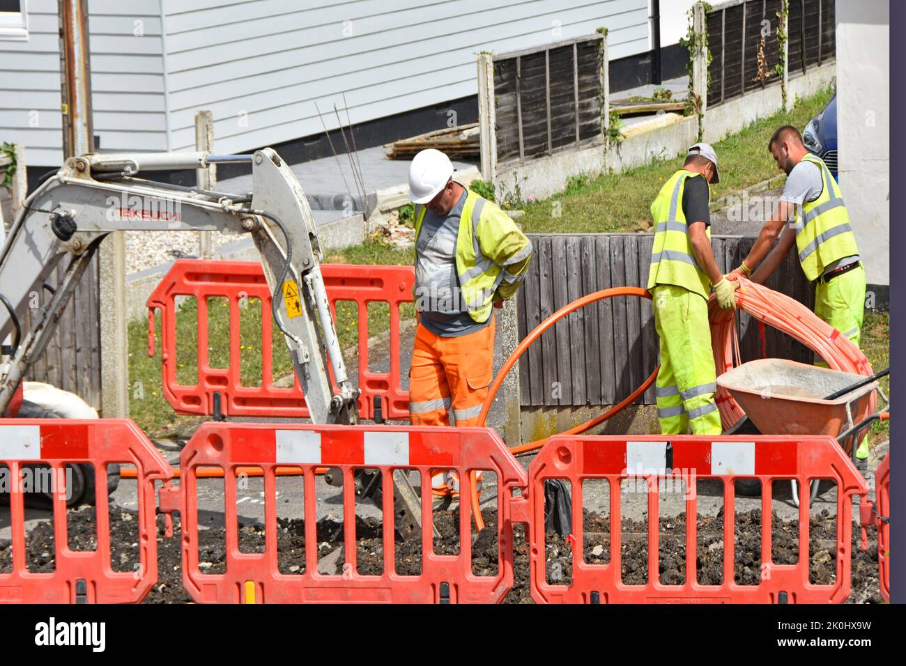 Group of workers installing coil of fibre optic high speed, broadband infrastructure cable into pavement trench outside homes in residential street UK Stock Photo