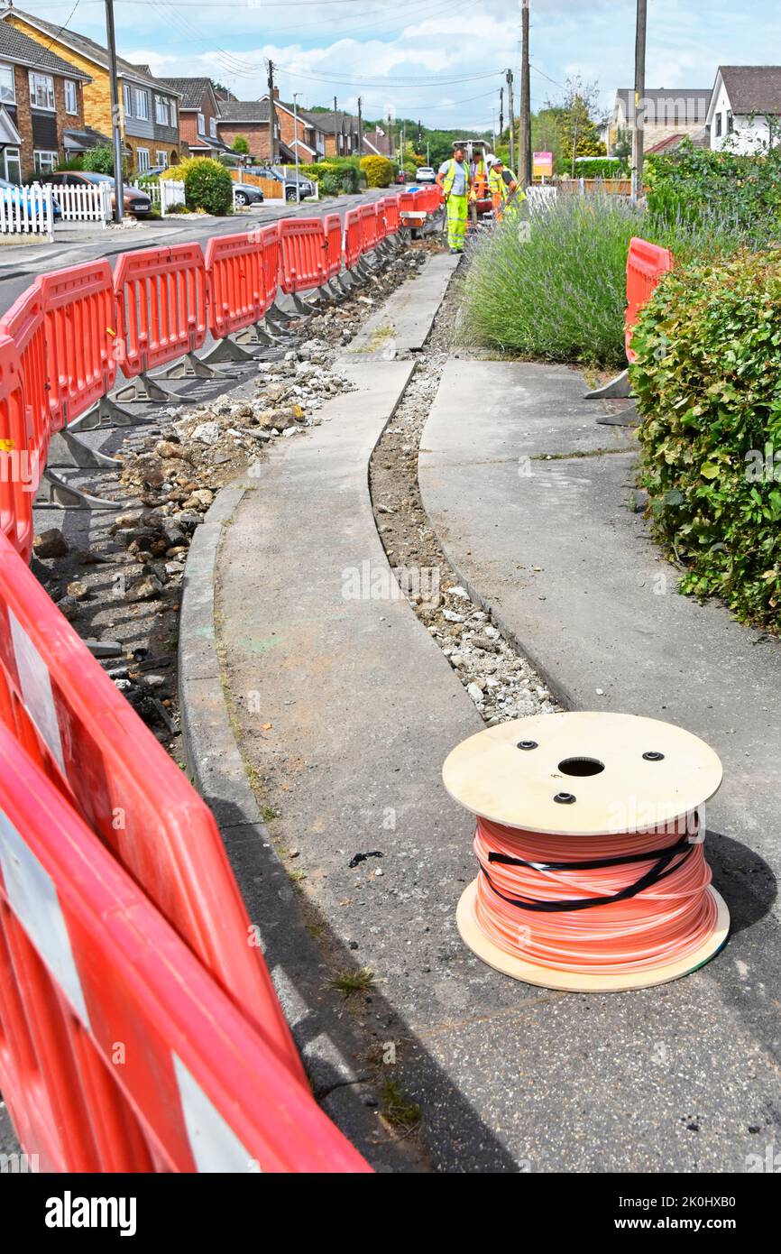 Workmen working on pavement reinstating fibre optic cable trench for new broadband infrastructure behind red interlocking safety barriers England UK Stock Photo