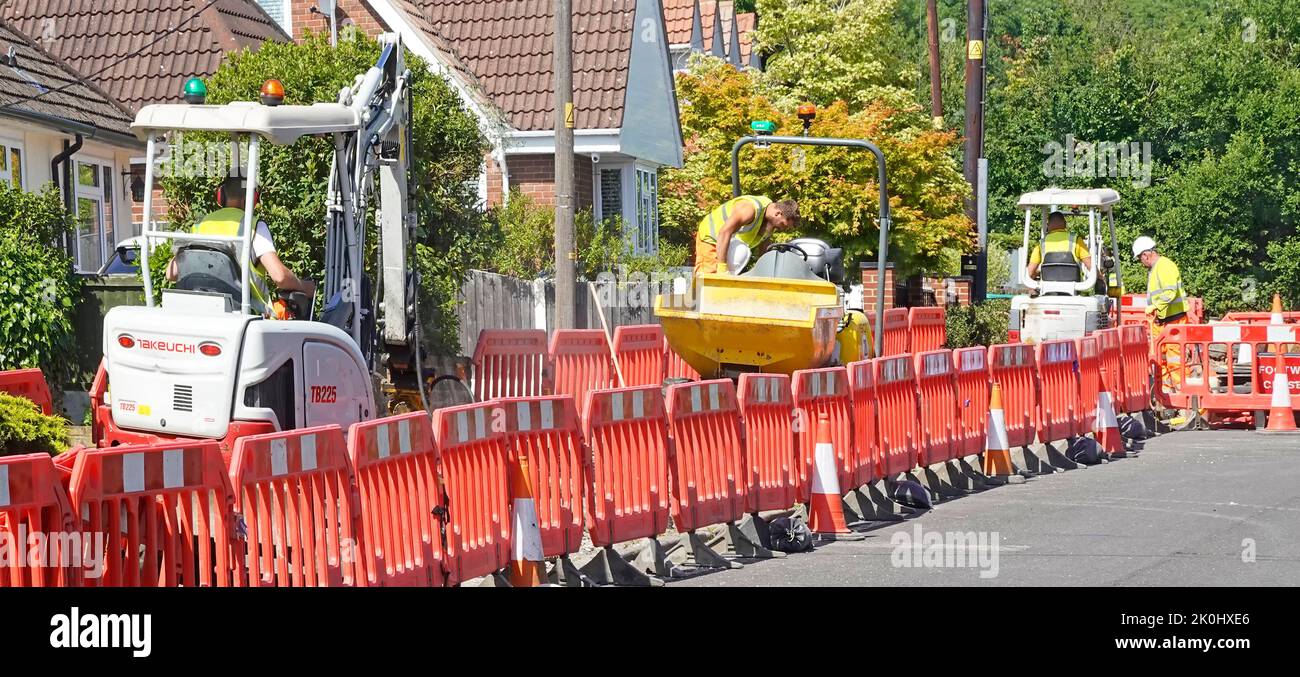 Workmen & excavating dumper machines working residential closed  pavement footway digging trench for fibre broadband cable infrastructure England UK Stock Photo