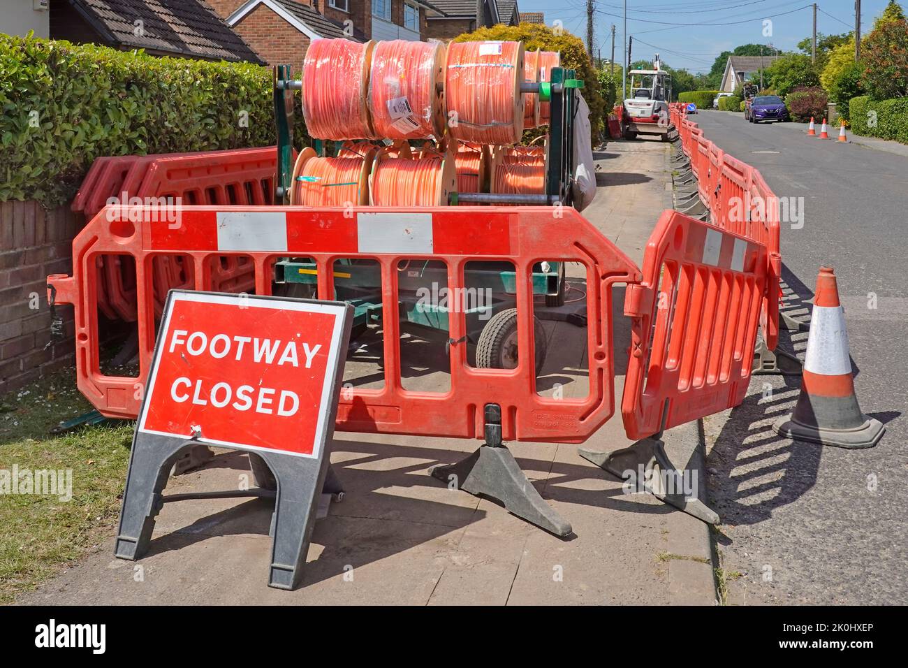 Footway Closed sign & housing pavement shut by red plastic road works safety barriers fast fibre broadband cable drums mini digger working England UK Stock Photo