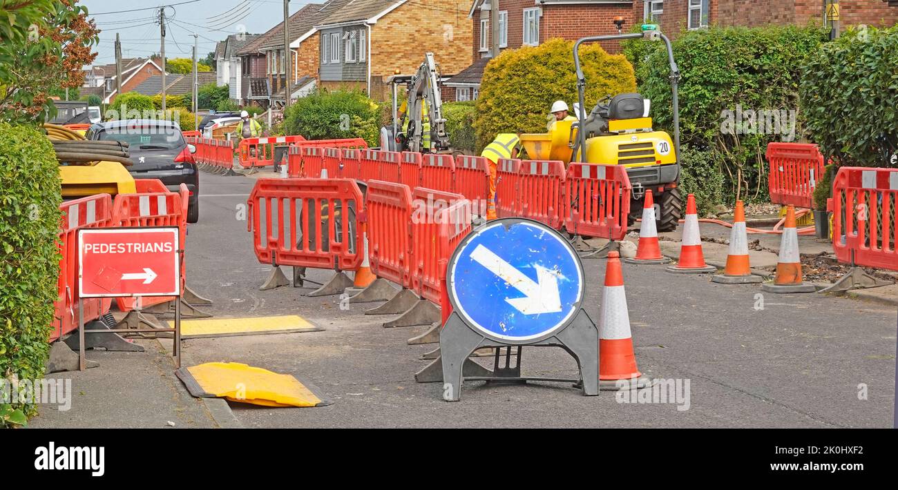 Red interlocking road works plastic safety barriers restricting cars & pedestrians access workers digging up pavement mini digger dumper machines UK Stock Photo