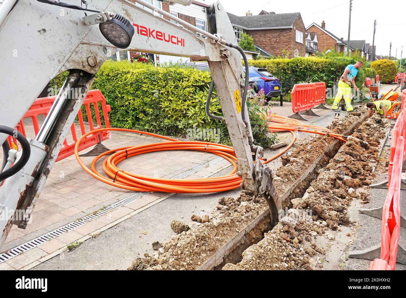 Close up mini digger excavator machine narrow trench channel in pavement for orange fibre optic broadband cable being positioned by workmen England UK Stock Photo