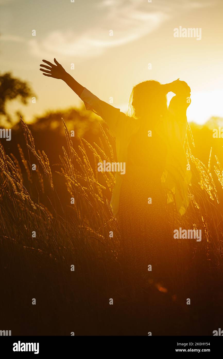 Smiling young blond woman standing amidst wheat field. Low angle. Dark shape against blinding light of a setting sun Stock Photo