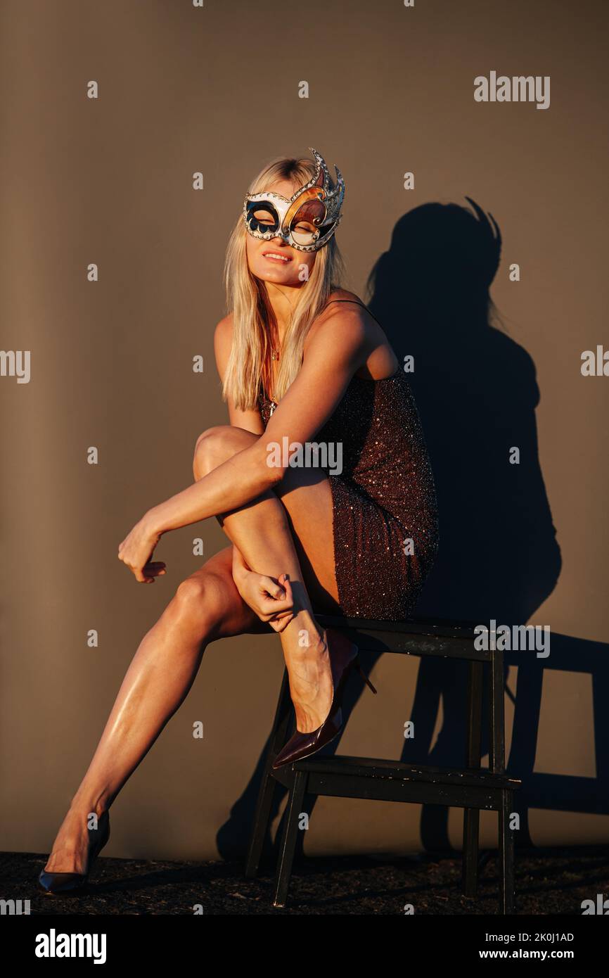 Taking a sunbath blond woman in a mask sitting on a stool, closing her eyes. Outdoors, sunlit. In front of brown background. Stock Photo
