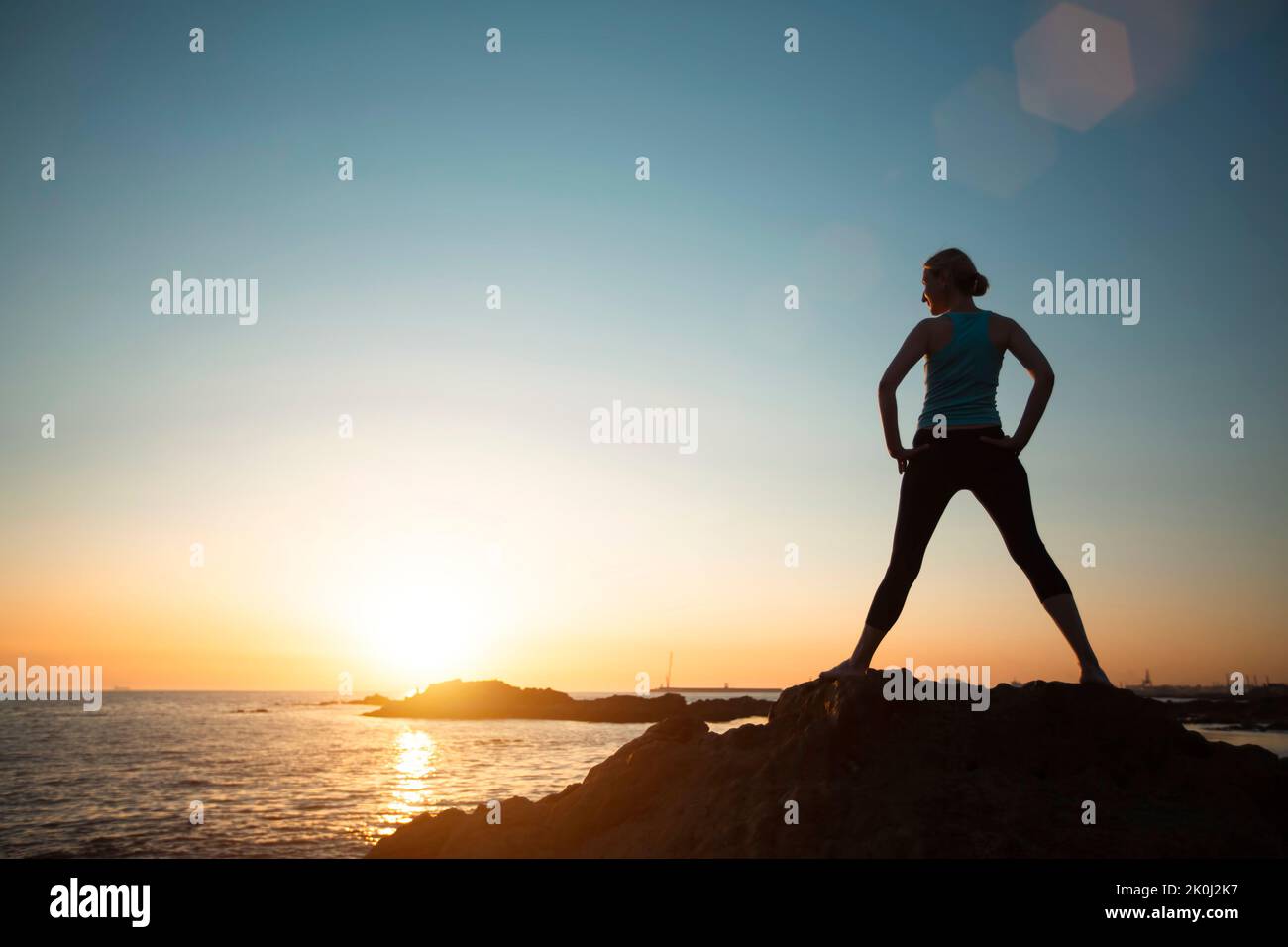 A woman does gymnastic yoga by the ocean in the setting sun. Stock Photo