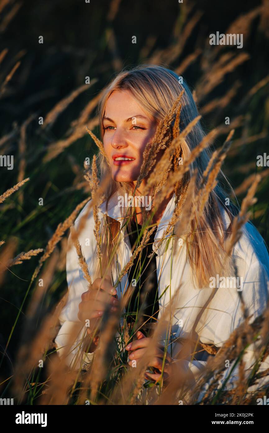 Young blond woman looking afar, standing amidst wheat field. Blurred background. Stock Photo