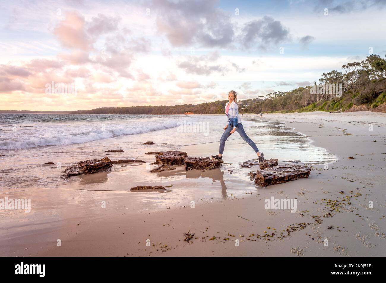 Pretty sky over the beach in the morning at Jervis Bay.  A woman steps between two rocks in the tide Stock Photo