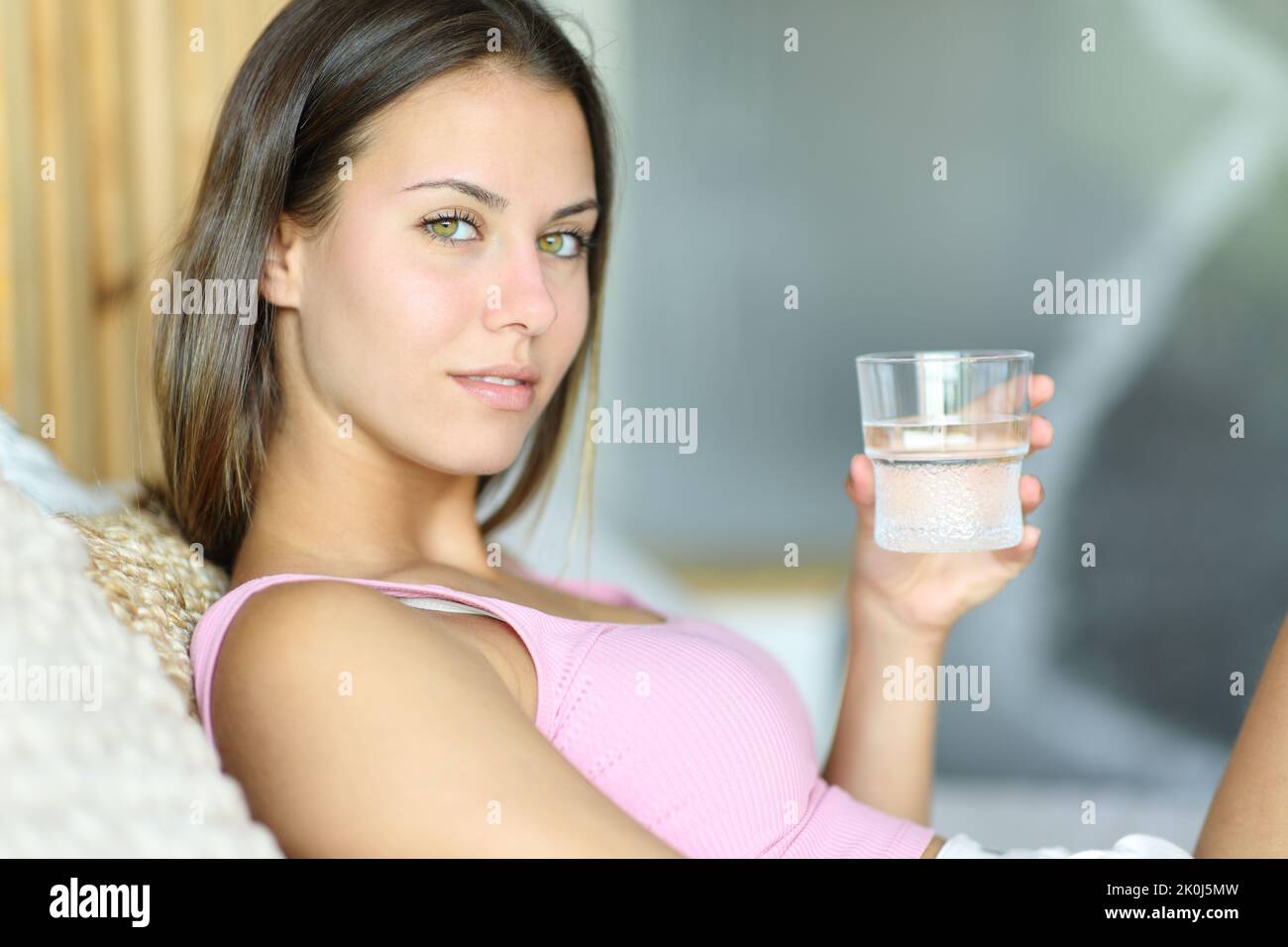 Beauty woman holding water glass looks at you sitting on a bed at home Stock Photo