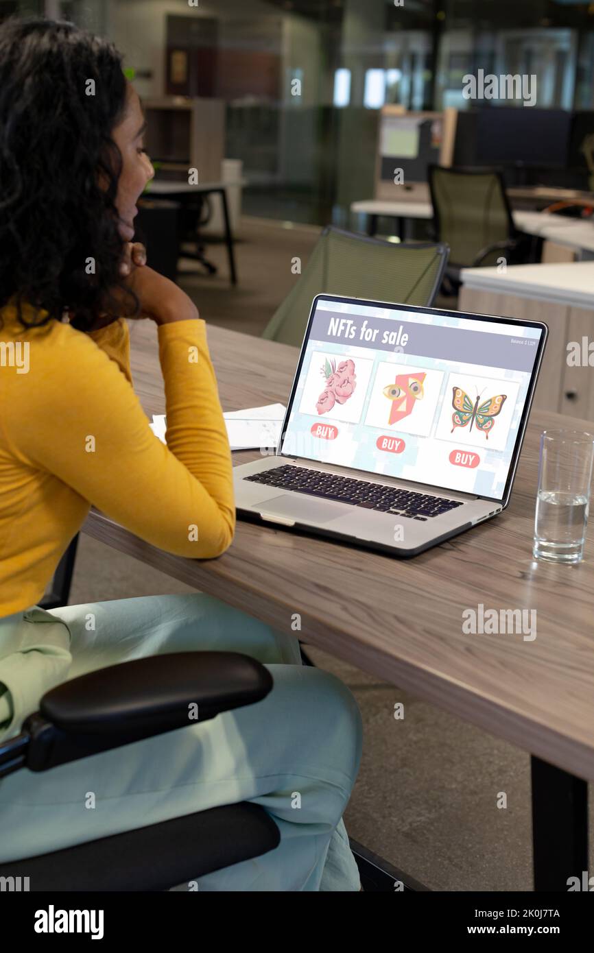Biracial businesswoman using laptop in office, with nfts for sale on screen. Business communication, non fungible tokens, cryptocurrency and digital i Stock Photo