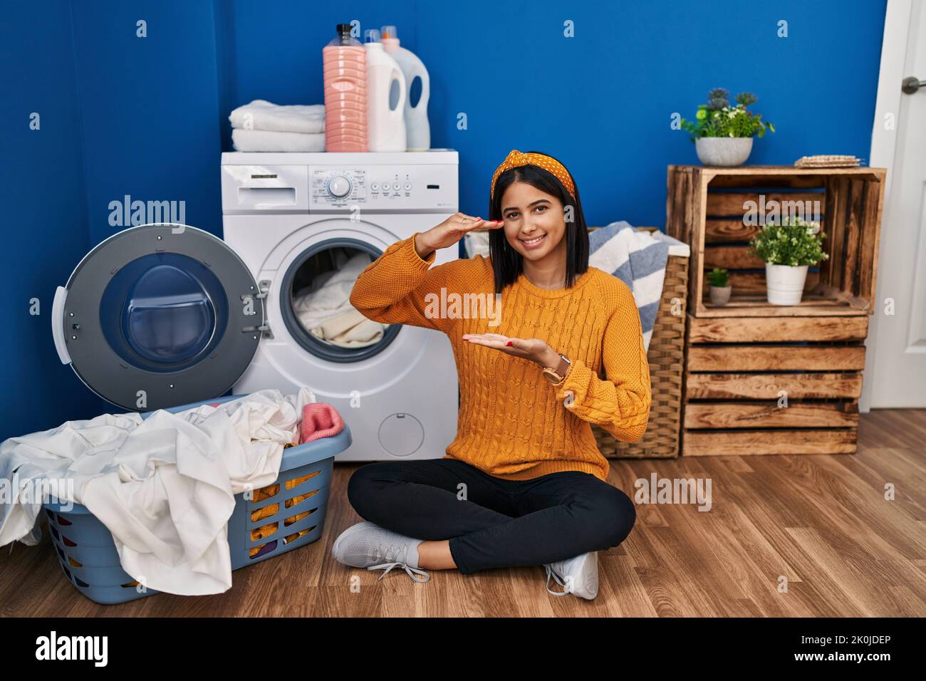 Young hispanic woman doing laundry gesturing with hands showing big and
