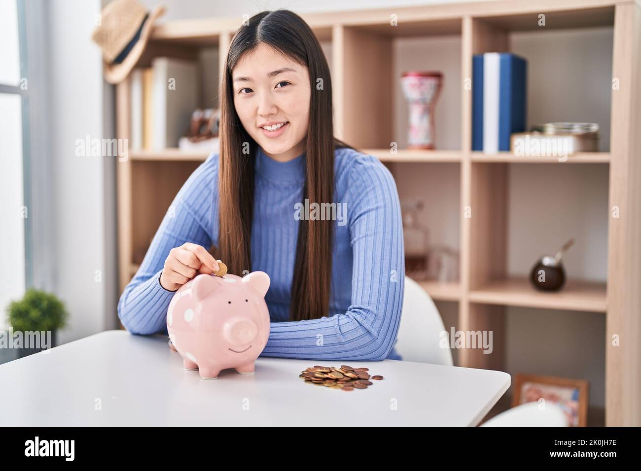 Chinese young woman putting coin in piggy bank looking positive and ...