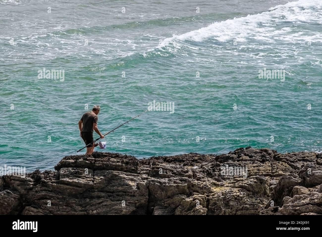 A holidaymaker carrying his fishing rod walking carefully on rocks at Crantock in Newquay in Cornwall in the UK. Stock Photo