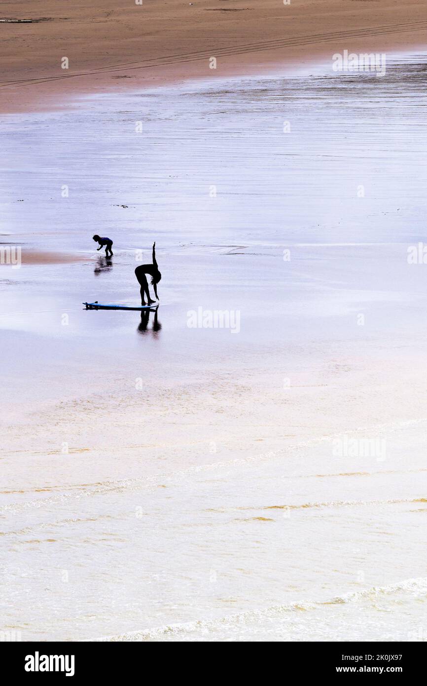 The figure of a surfer seen in silhouette stretching before a surfing session on Crantock Beach; in Newquay in Cornwall in the UK. Stock Photo