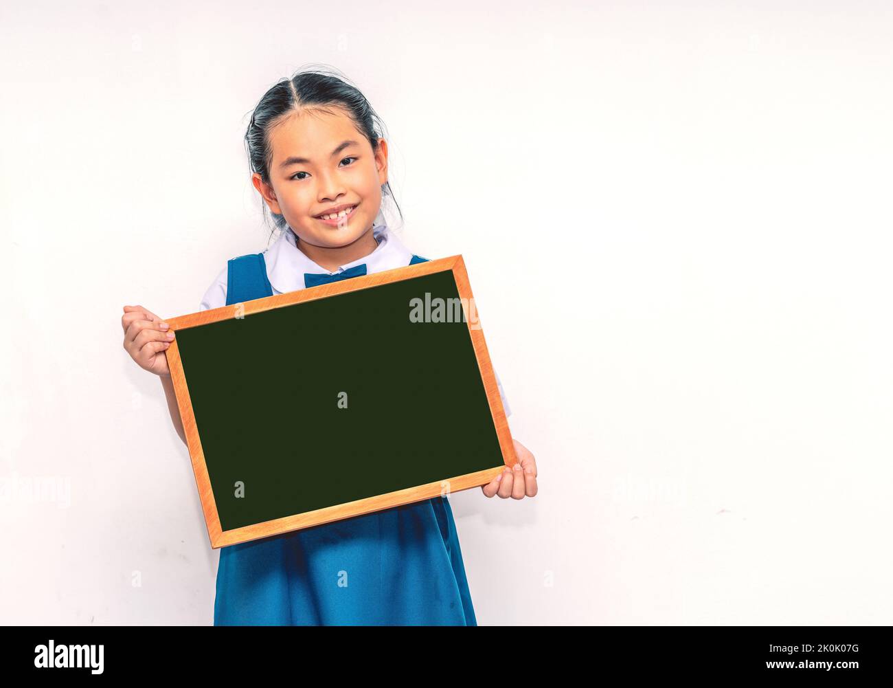 Portrait Asian student child in identified school uniform with smiling face, holding small blackboard or chalkboard, isolated image on white backgroun Stock Photo