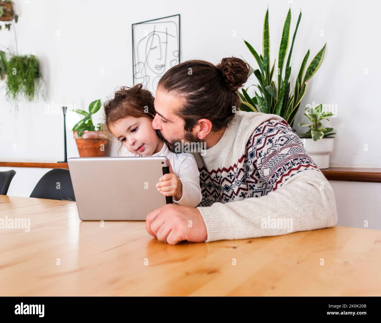 Bearded male in sweater sitting at table and kissing on cheek daughter using tablet in light apartment with potted plants Stock Photo