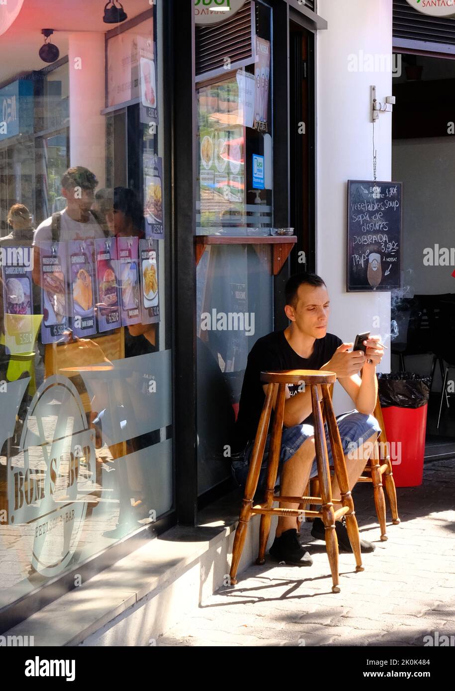 Man sitting, smoking and using mobile phone outside cafe in Camara do Lobos, Madeira, Portugal Stock Photo