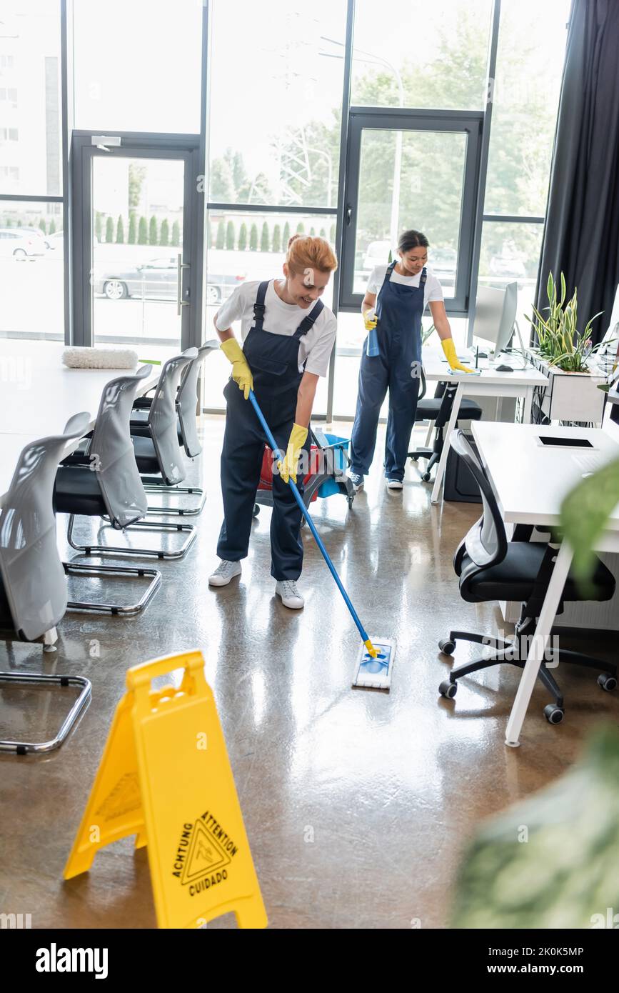 happy woman washing floor with mop near caution board and mixed race