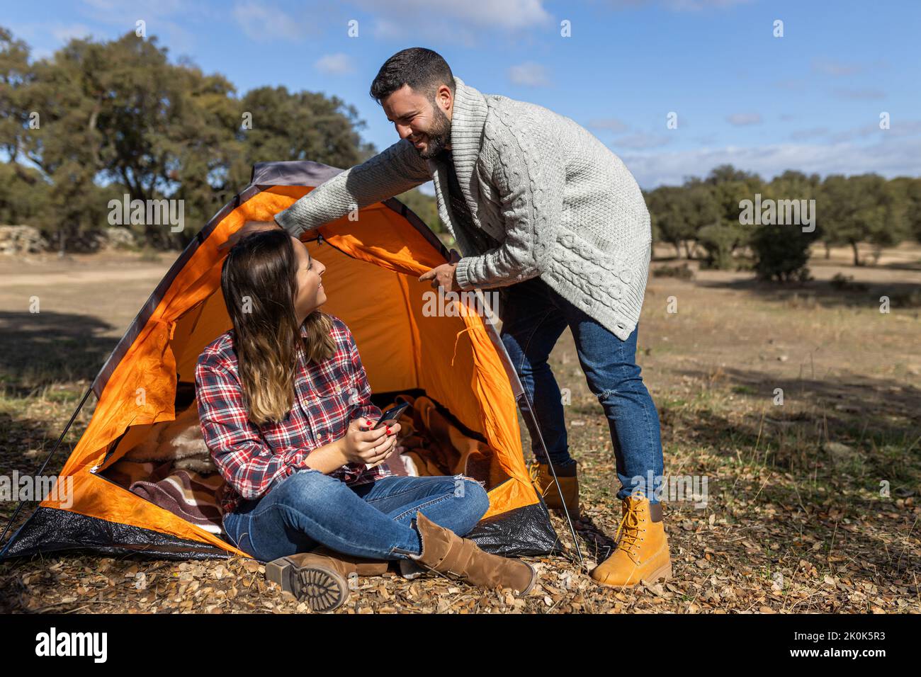 Young female browsing on mobile phone while boyfriend prepares camping tent in forest Stock Photo