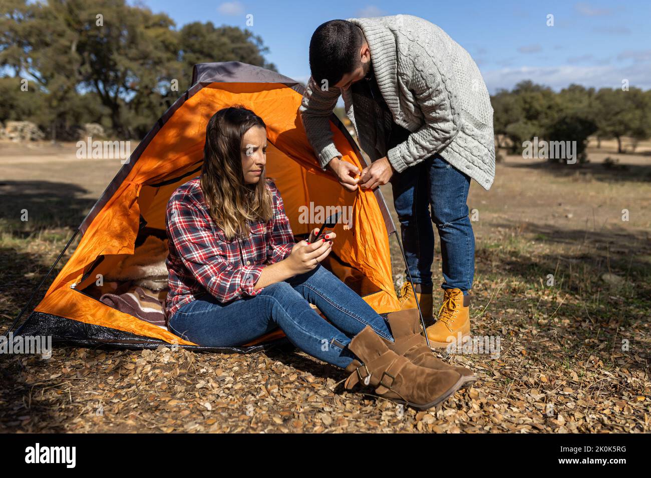 Young female browsing on mobile phone while boyfriend prepares camping tent in forest Stock Photo