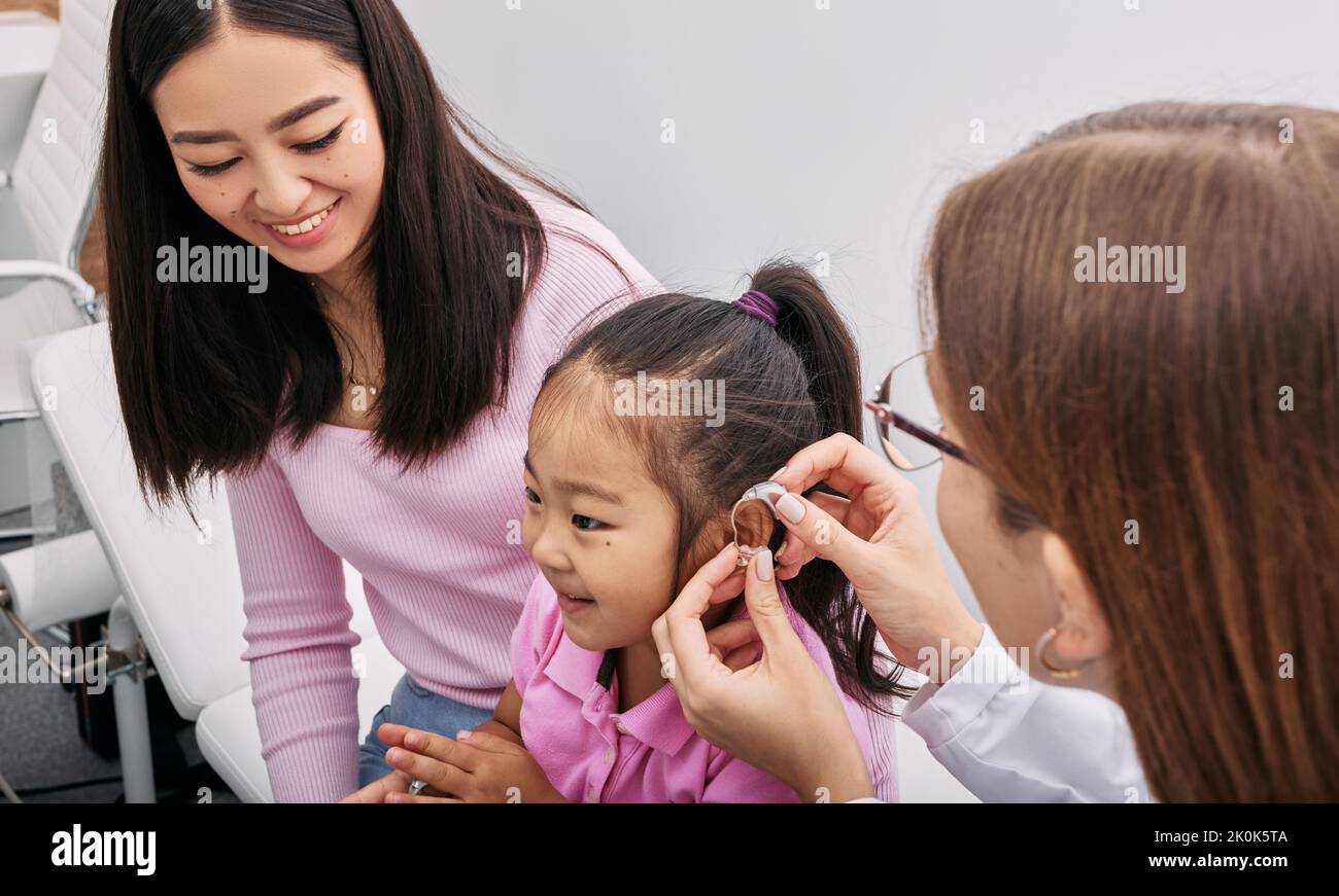 Audiologist fits hearing aid on Asian little girl's ear while visit in hearing clinic with mother. Hearing solutions for children Stock Photo