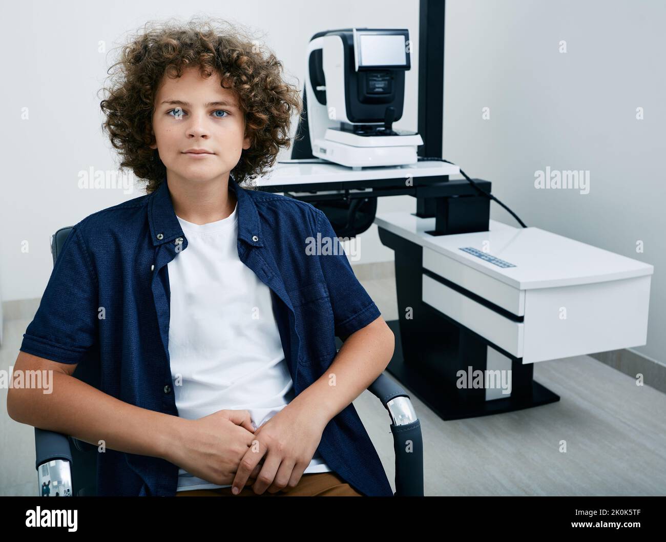 Cute boy sitting in ophthalmology clinic near ophthalmic equipment for eye examination. Ophthalmologist will check visual acuity of child using autore Stock Photo