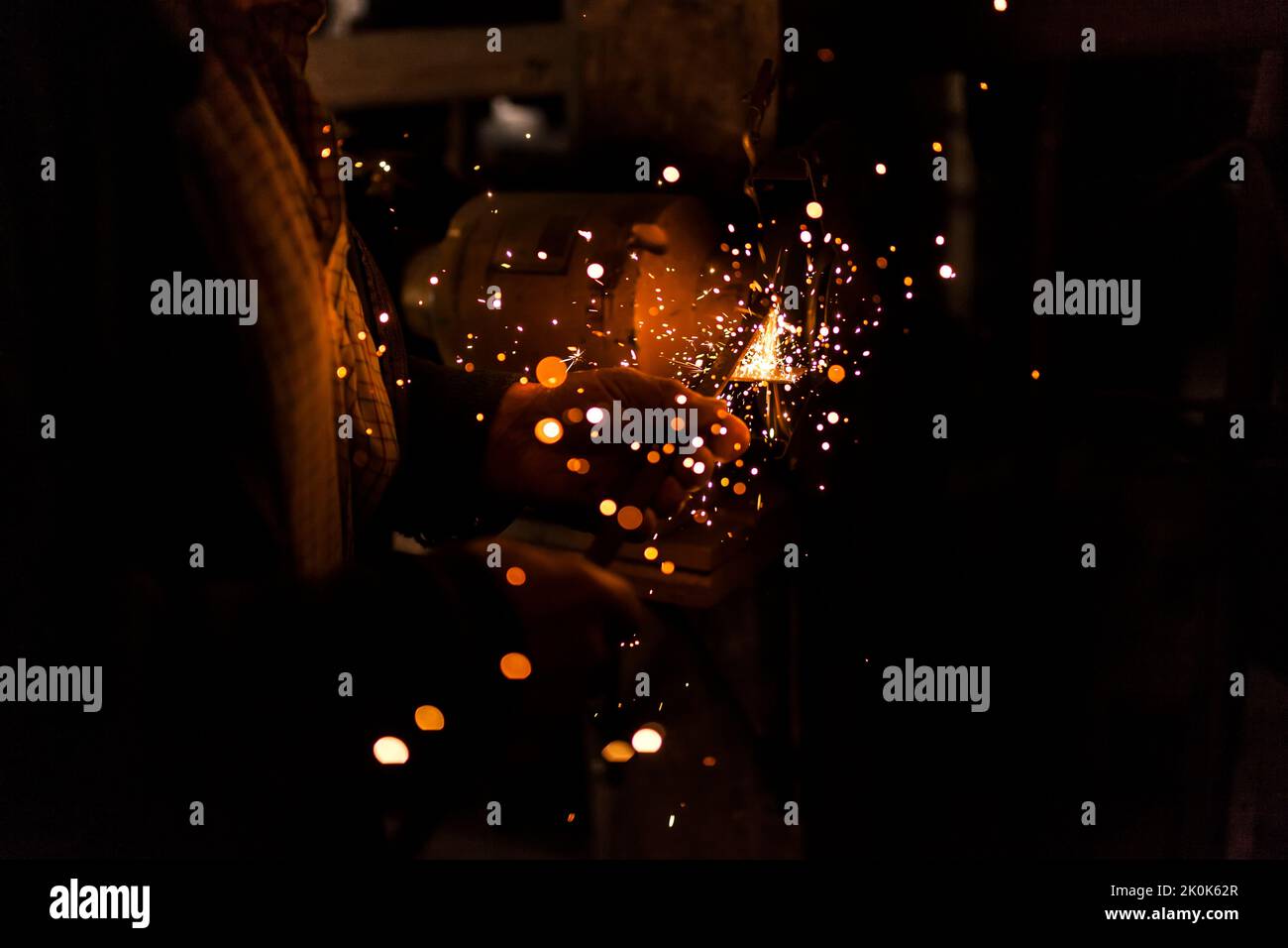 Unrecognizable elderly craftsman using bench grinder to shape metal part while working in dark workshop Stock Photo