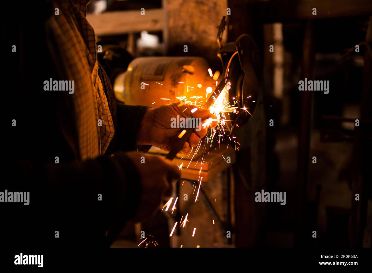 Unrecognizable elderly craftsman using bench grinder to shape metal part while working in dark workshop Stock Photo