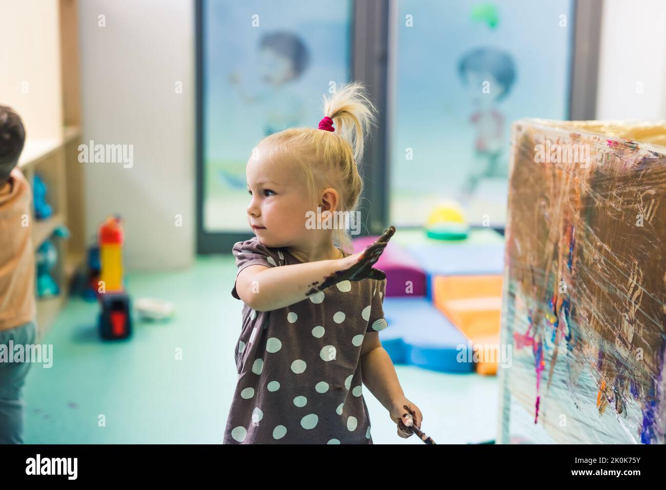 Cling film painting. Little caucasian girl toddler painting with her arms on a cling film wrapped all the way round the wooden shelf unit. Creative activity for kids development at the nursery school. High quality photo Stock Photo